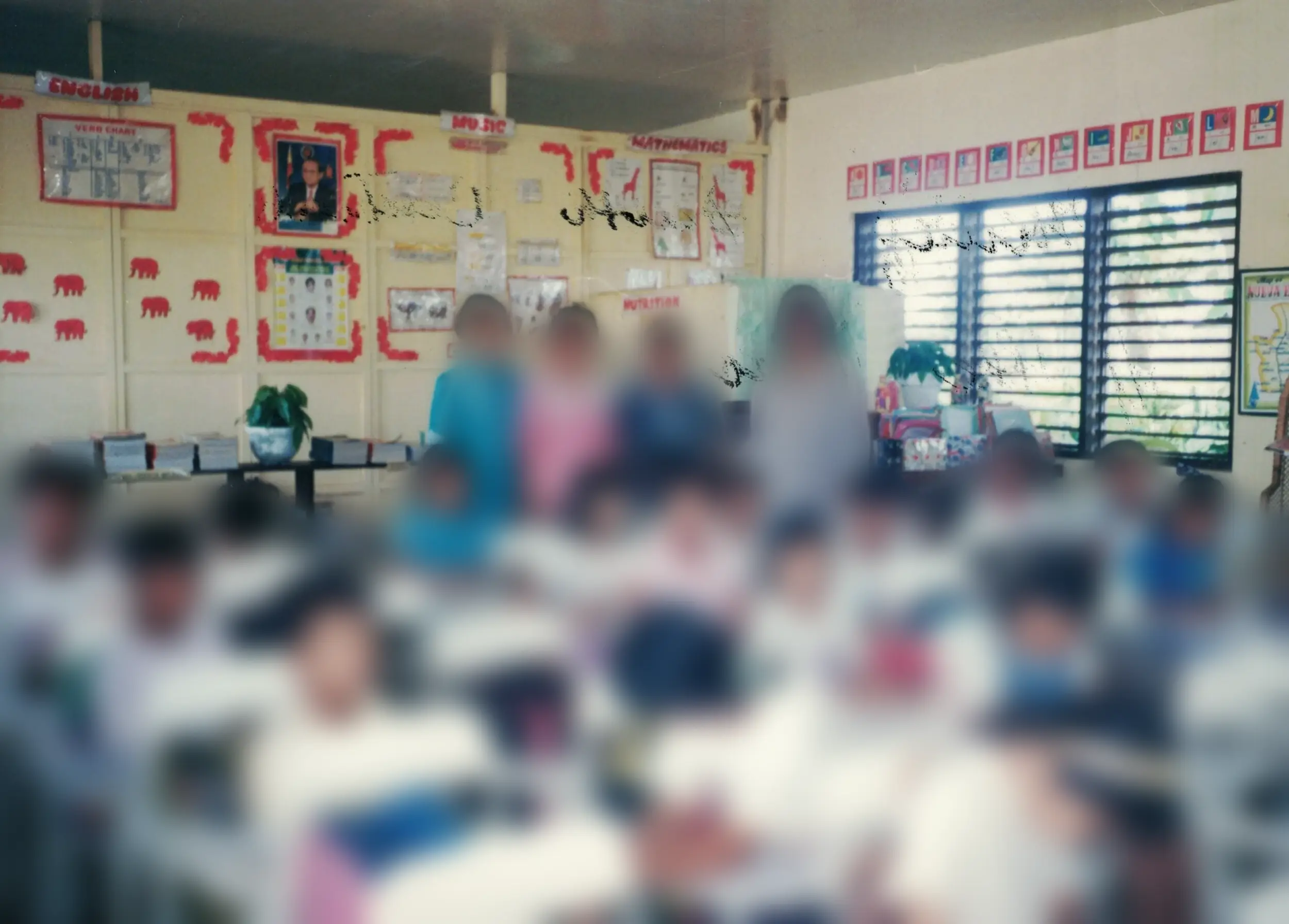 Scanned photo of a grade-school classroom: children seated at white desks, four women standing at the back beneath English, Music, and Mathematics posters.